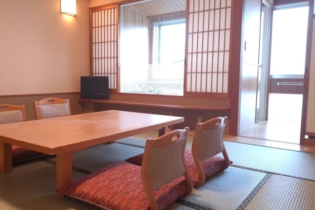 A traditional Japanese room with tatami flooring, a low table, four chairs, and a television.