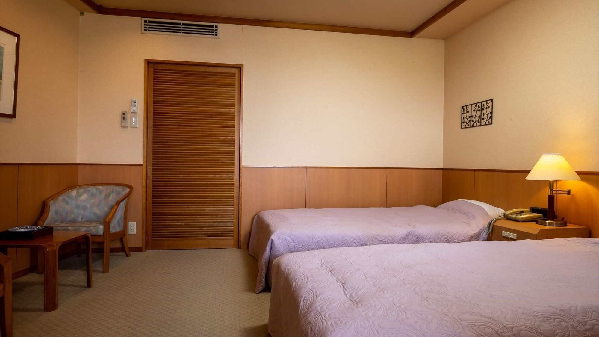 A hotel room with two beds, a chair, a desk, a lamp, and a wall-mounted phone.