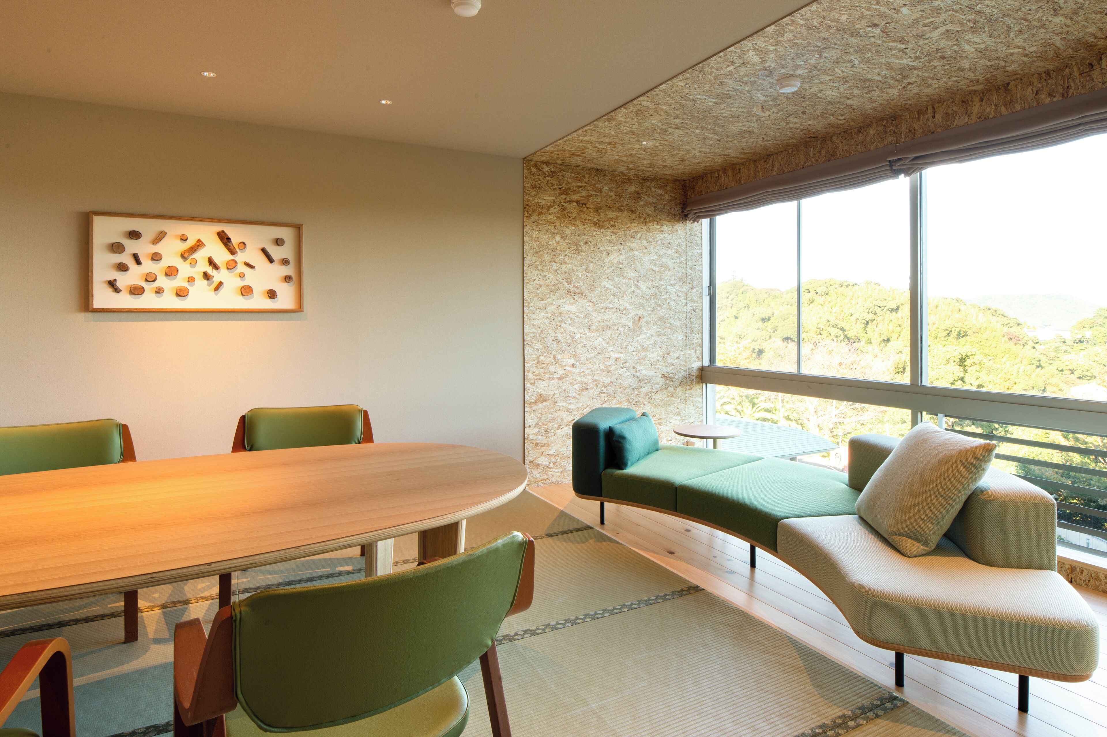 A modern meeting room with a wooden table, green chairs, a sofa, and a large window offering a view of greenery.