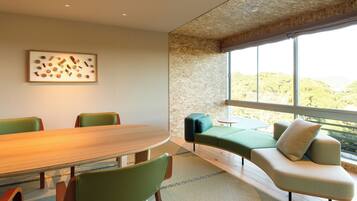A modern meeting room with a wooden table, green chairs, a sofa, and a large window offering a view of greenery.