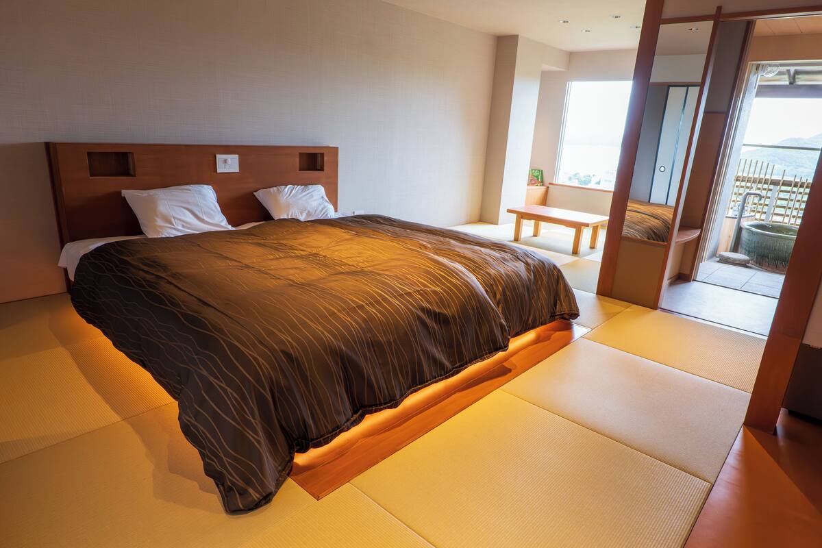 A modern hotel room with a large bed, a wooden headboard, and a view of the outdoors through a sliding door.