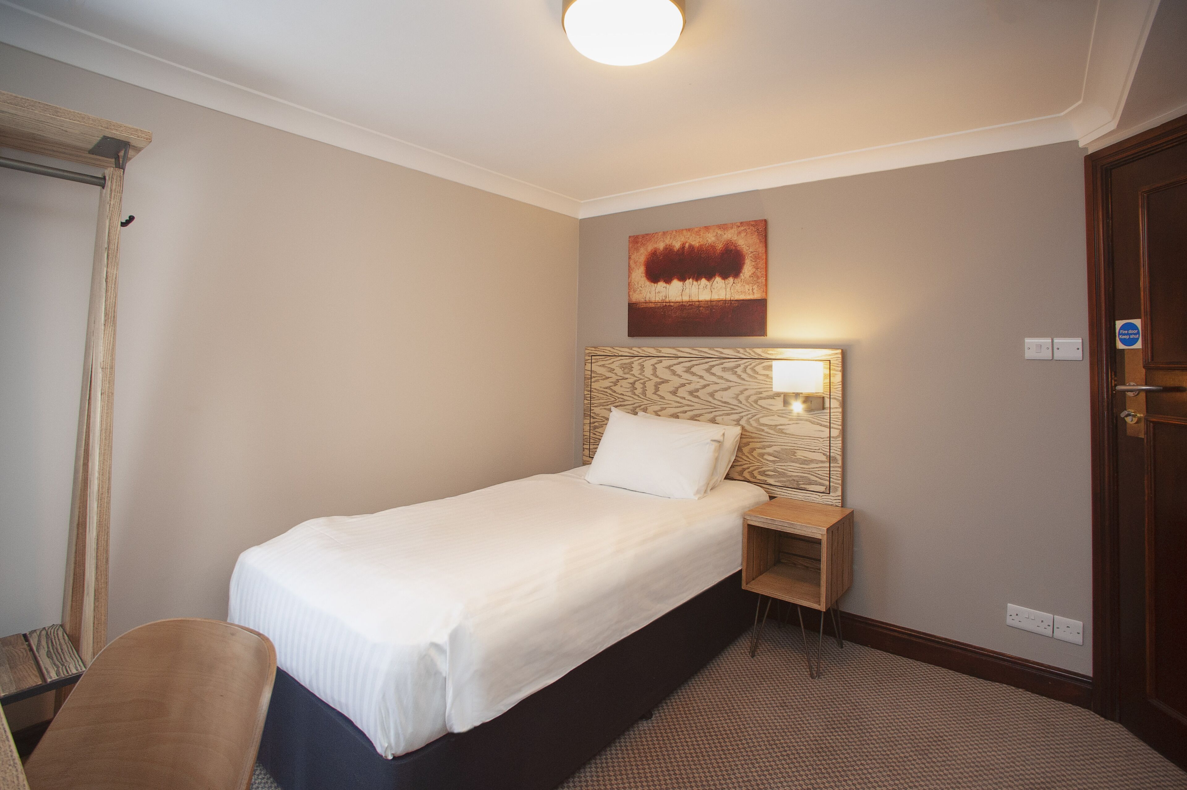 Una habitación de hotel con una cama individual, una mesita de noche, una lámpara de pared y una puerta.