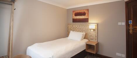 A hotel room with a single bed, a bedside table, a nightstand, a wall-mounted lamp, and a door.