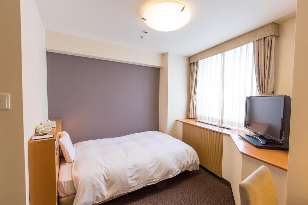 Une chambre d’hôtel avec un lit simple, un bureau avec une télévision, une fenêtre avec des rideaux et une petite table de chevet avec un téléphone.