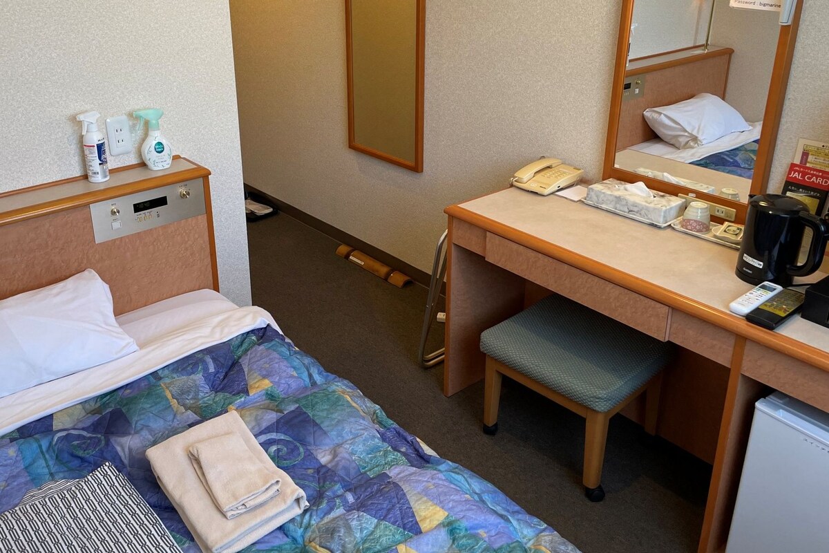 A hotel room with a bed, a desk, a chair, a small refrigerator, a mirror, a telephone, and toiletries on the nightstand.