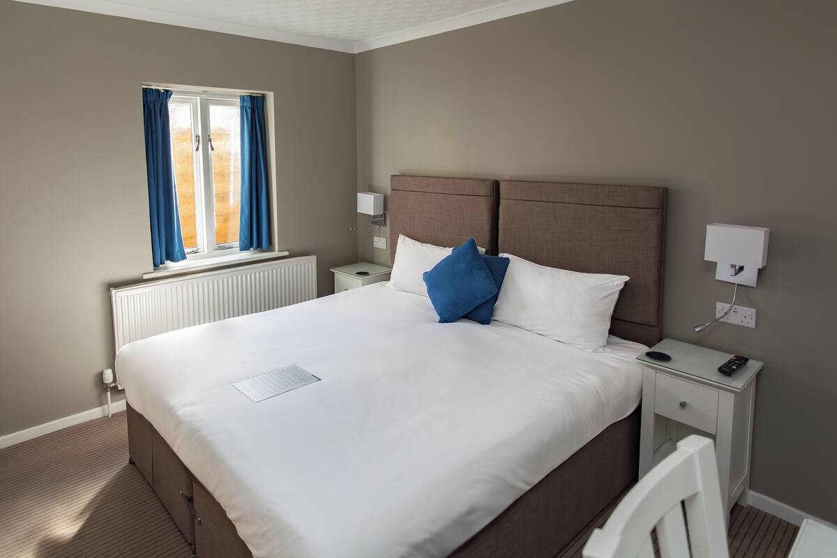 A neatly made bed with white and blue pillows, a bedside table with a remote control, and a window with blue curtains.
