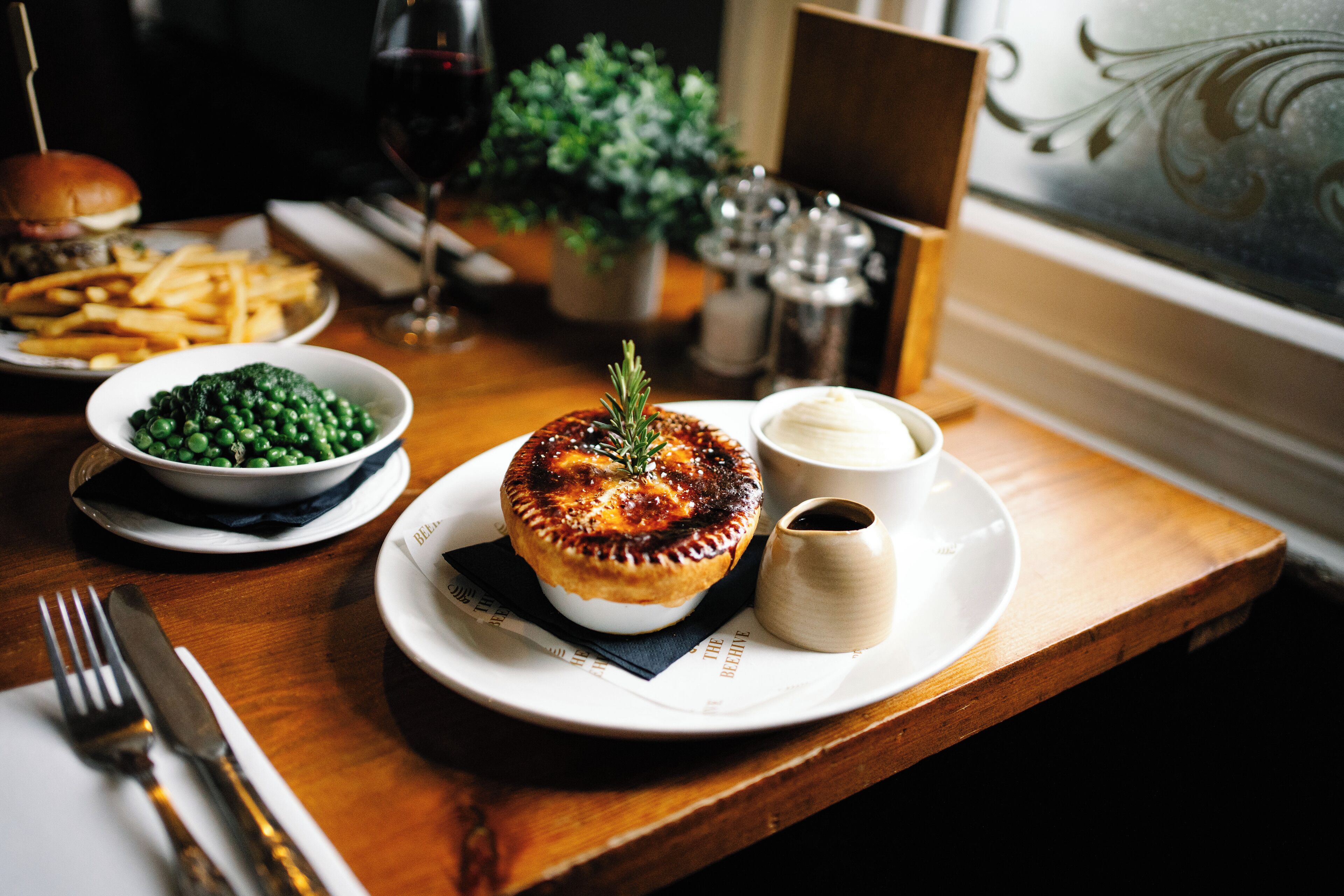A dining table with a meal consisting of a pie, peas, fries, and a burger.