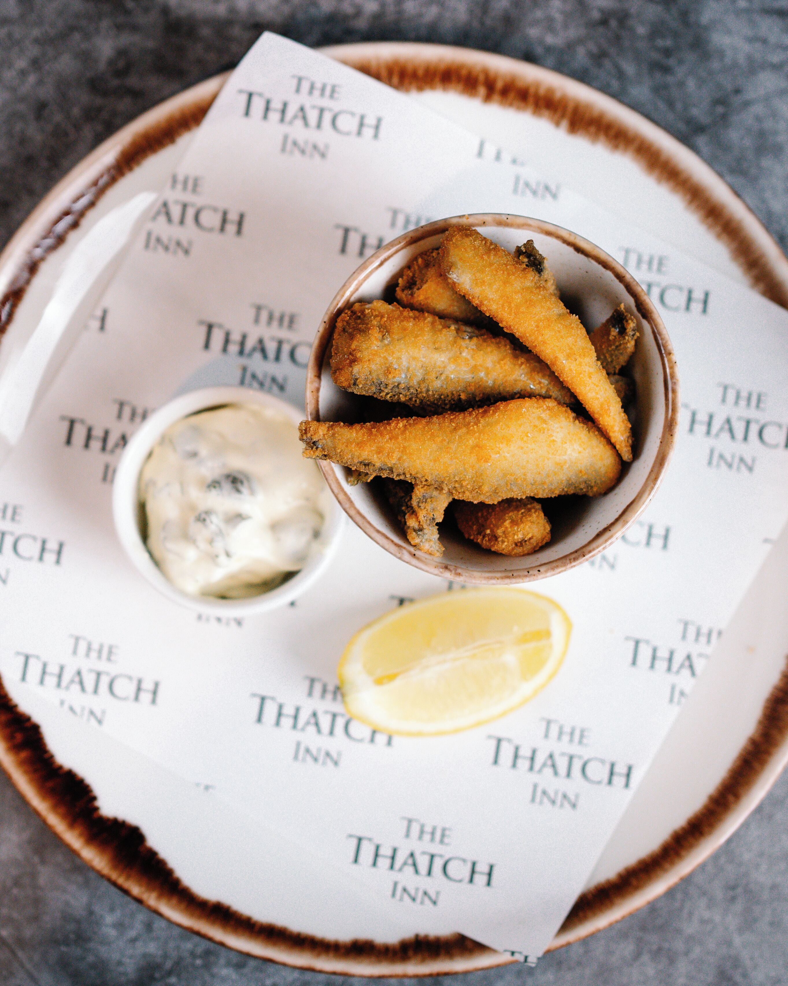 Un plato de pescado frito con un gajo de limón y un pequeño recipiente de salsa tártara, colocado sobre un menú de The Hatch Inn.