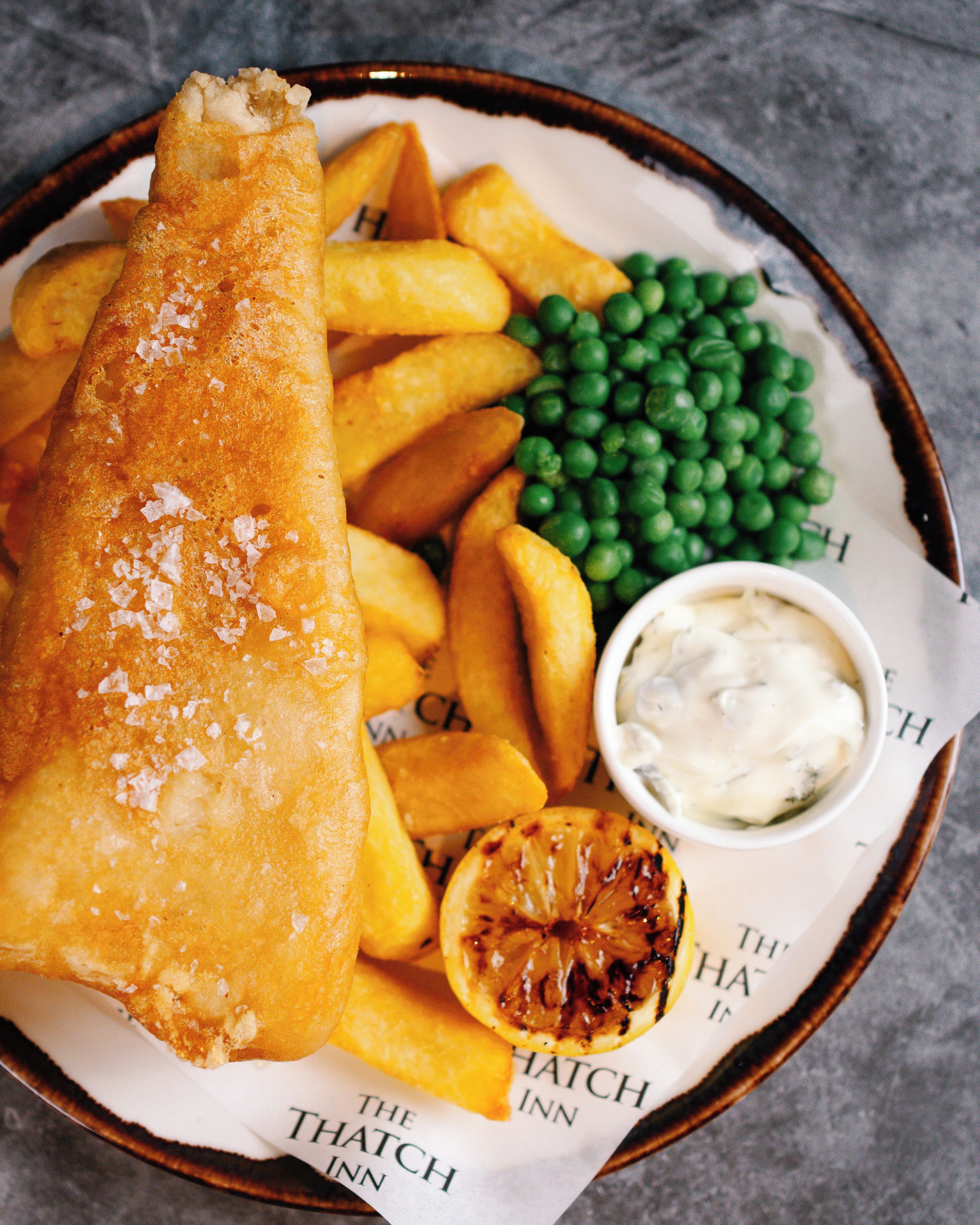A plate of fish and chips, peas, and a slice of grilled lemon.