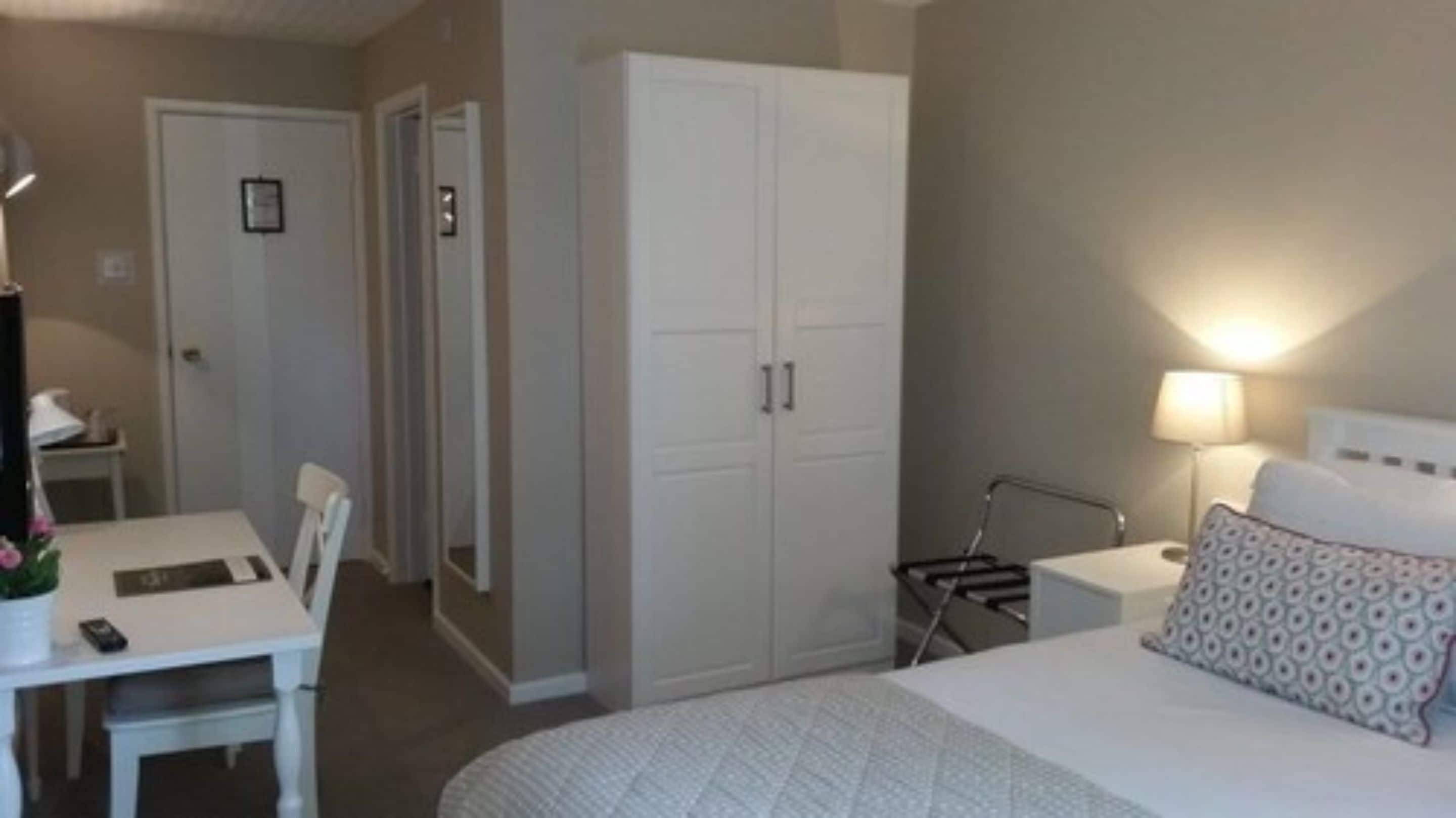 Una habitación de hotel con cama, escritorio, silla, armario y mesita de noche con lámpara.