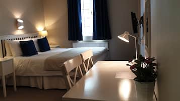 A hotel room with a bed, bedside table, chair, desk, lamp, and a potted plant.