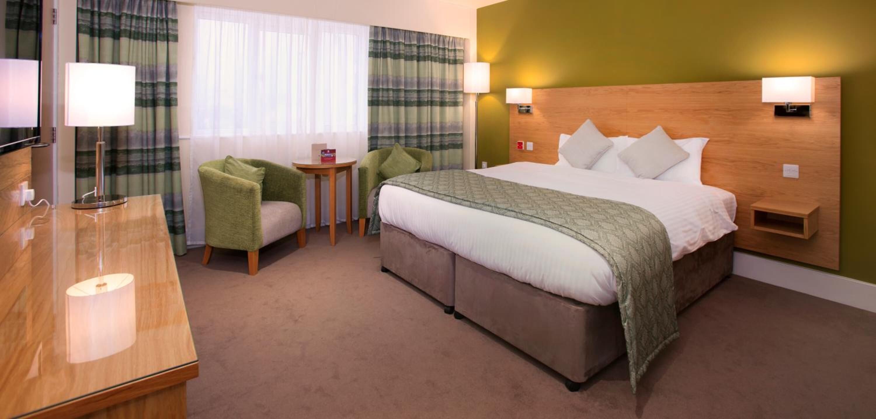 A hotel room with a bed, two armchairs, a small table, a bedside table, and a lamp.