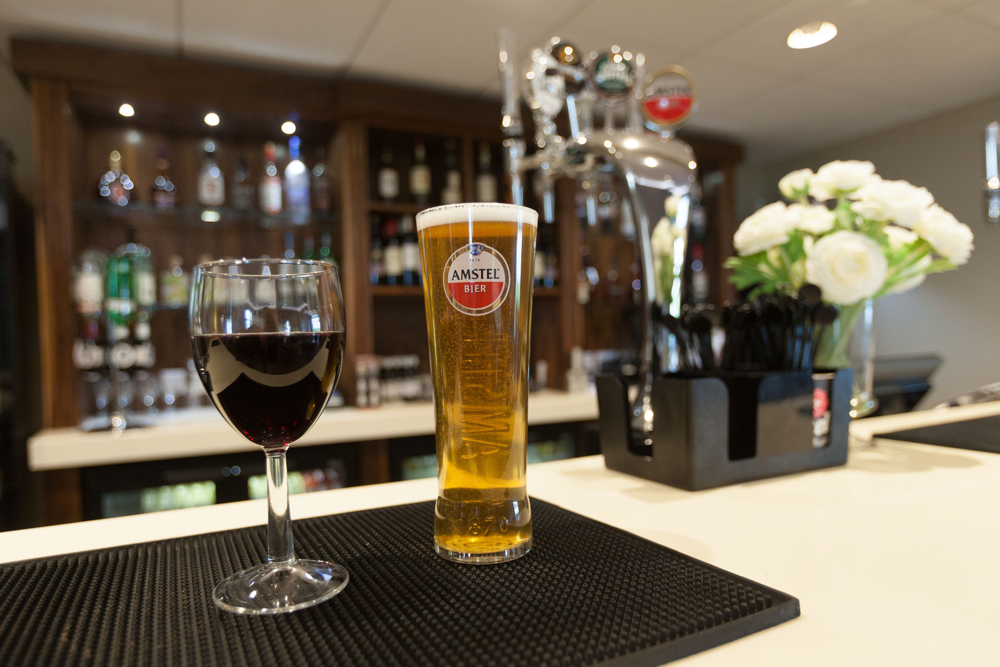 A bar area with a glass of red wine and a tall beer glass labeled "AMSTEL BIER" on a black mat.