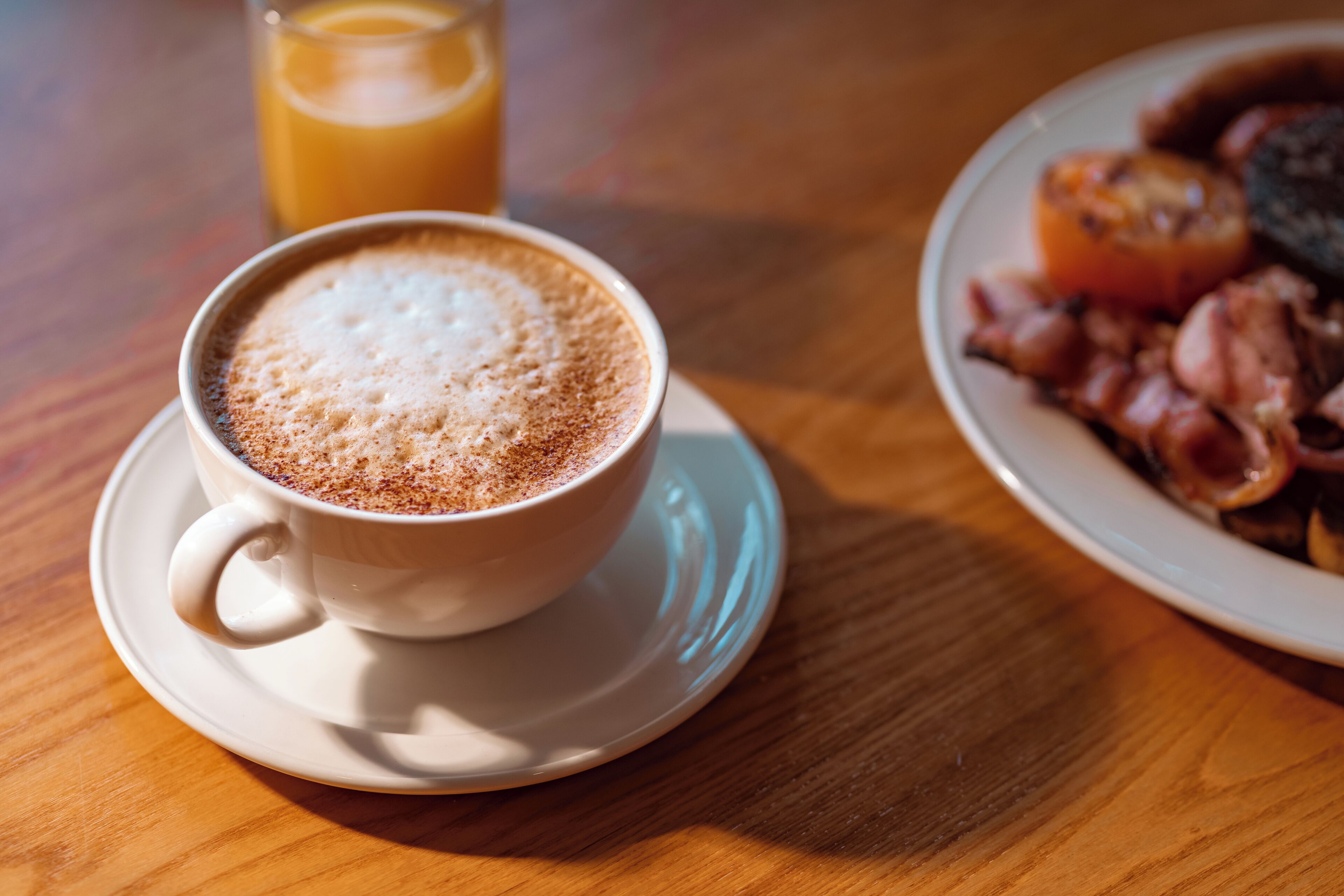 A cup of coffee with frothy milk on a saucer, a glass of orange juice, and a plate of bacon and eggs.