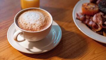 A cup of coffee with frothy milk on a saucer, a glass of orange juice, and a plate of bacon and eggs.