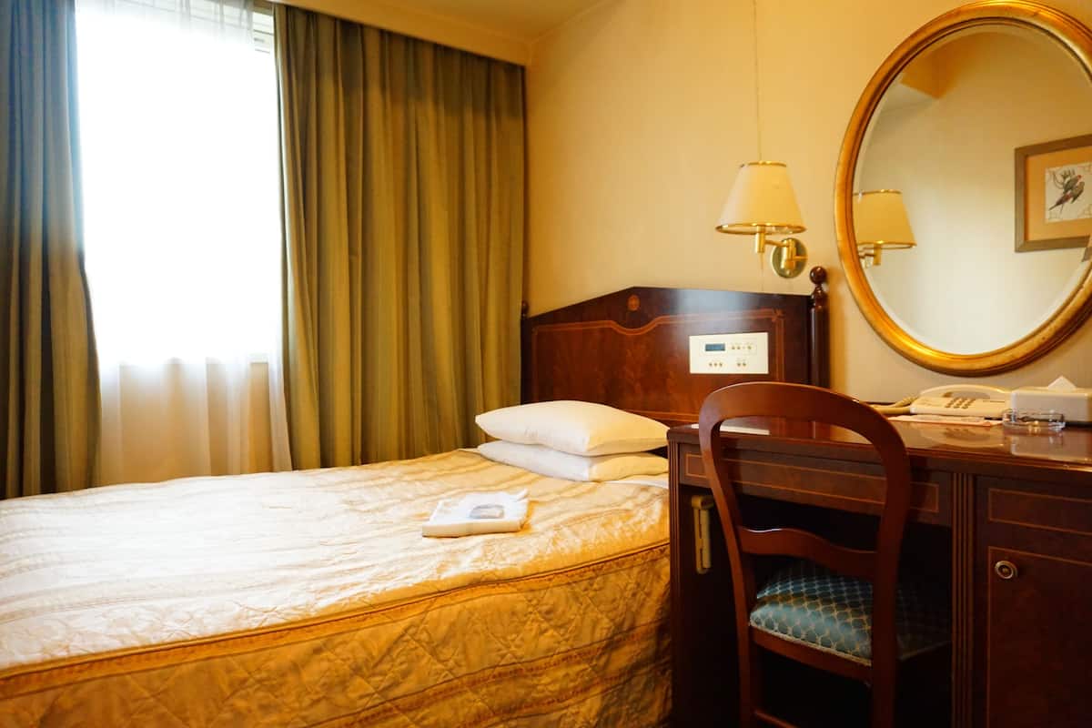 A hotel room with a bed, a wooden nightstand, a chair, a desk, a mirror, and a window with curtains.