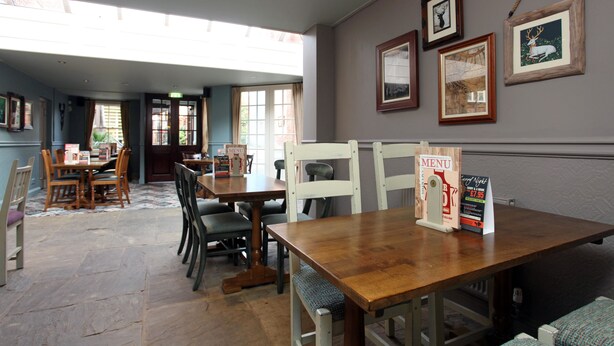 A restaurant with wooden tables and chairs, framed pictures on the wall, and a menu stand. - Original White Hart, Ringwood by Marston's Inns (Ringwood)