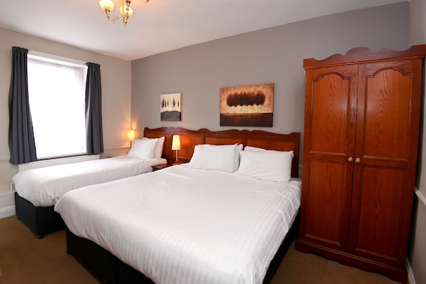 A hotel room with two beds, a wardrobe, a window with curtains, and two wall-mounted lamps. - Original White Hart, Ringwood by Marston's Inns (Ringwood)