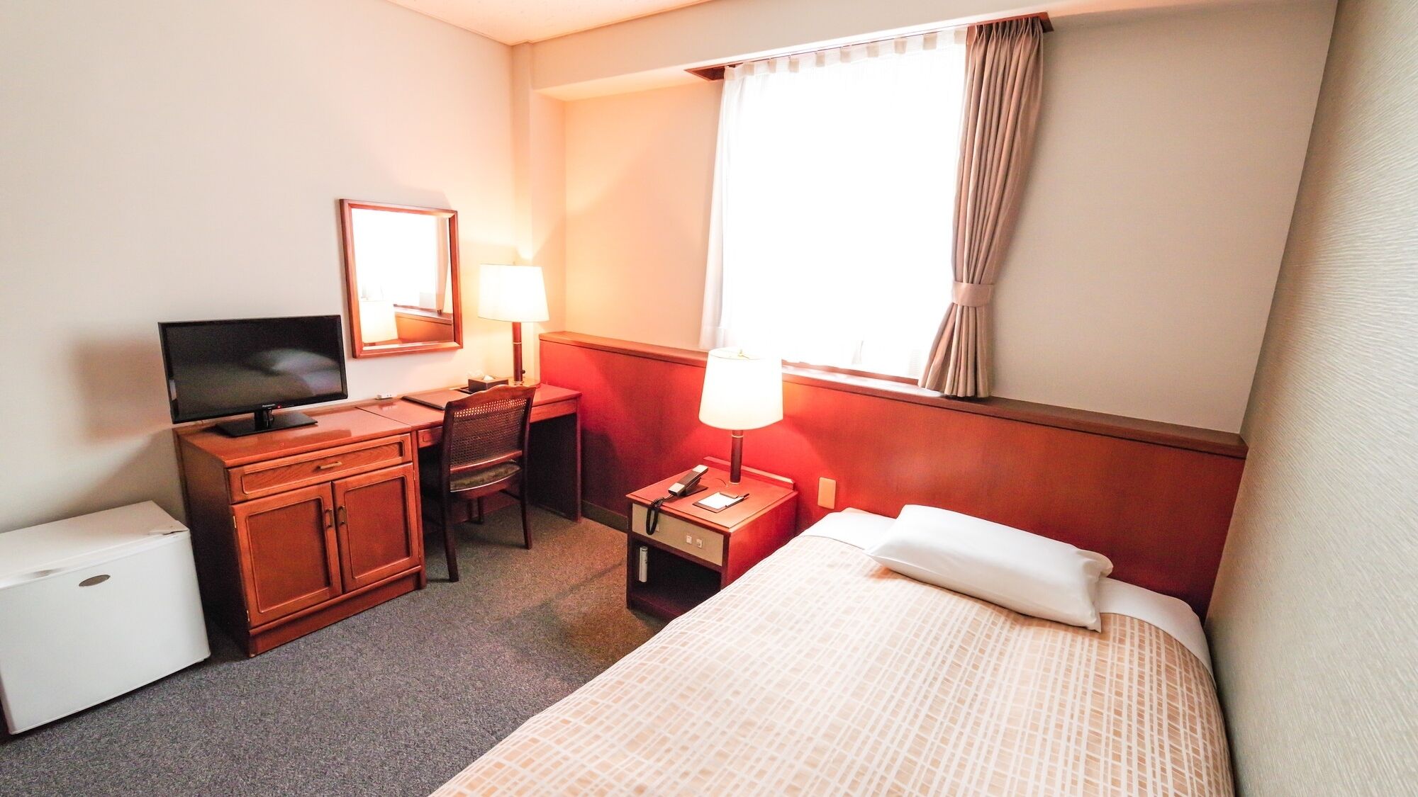 A hotel room with a bed, a desk with a computer and lamp, a TV, a small fridge, and a window with curtains.