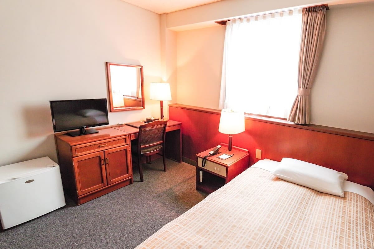 A hotel room with a bed, a desk with a computer and lamp, a TV, a small fridge, and a window with curtains.