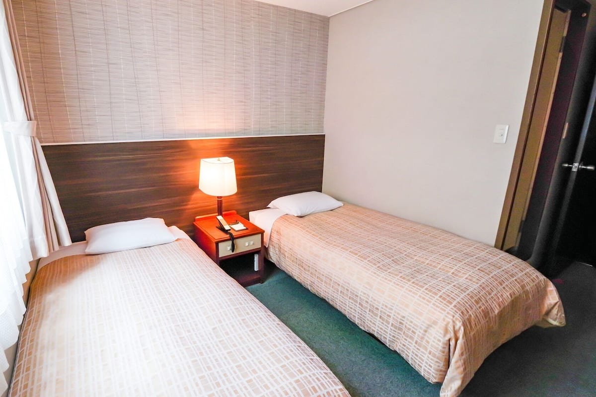 A hotel room with two single beds, a nightstand with a lamp, and a door to the right.