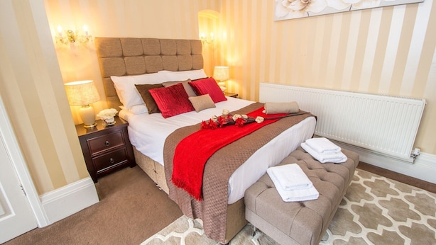 A hotel room with a large bed, a bedside table with a lamp, a radiator, and a wall with a floral painting. - Norman Hurst (Sale)