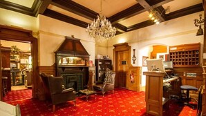 A room with a red patterned carpet, a chandelier, a fireplace, a wooden desk, and a bookshelf. - Norman Hurst (Sale)