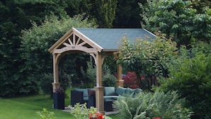 A wooden gazebo with a green roof, surrounded by lush greenery and a variety of plants. - Norman Hurst (Sale)