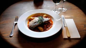 A plate of fish with vegetables and sauce, a fork, a knife, and two wine glasses on a wooden table.