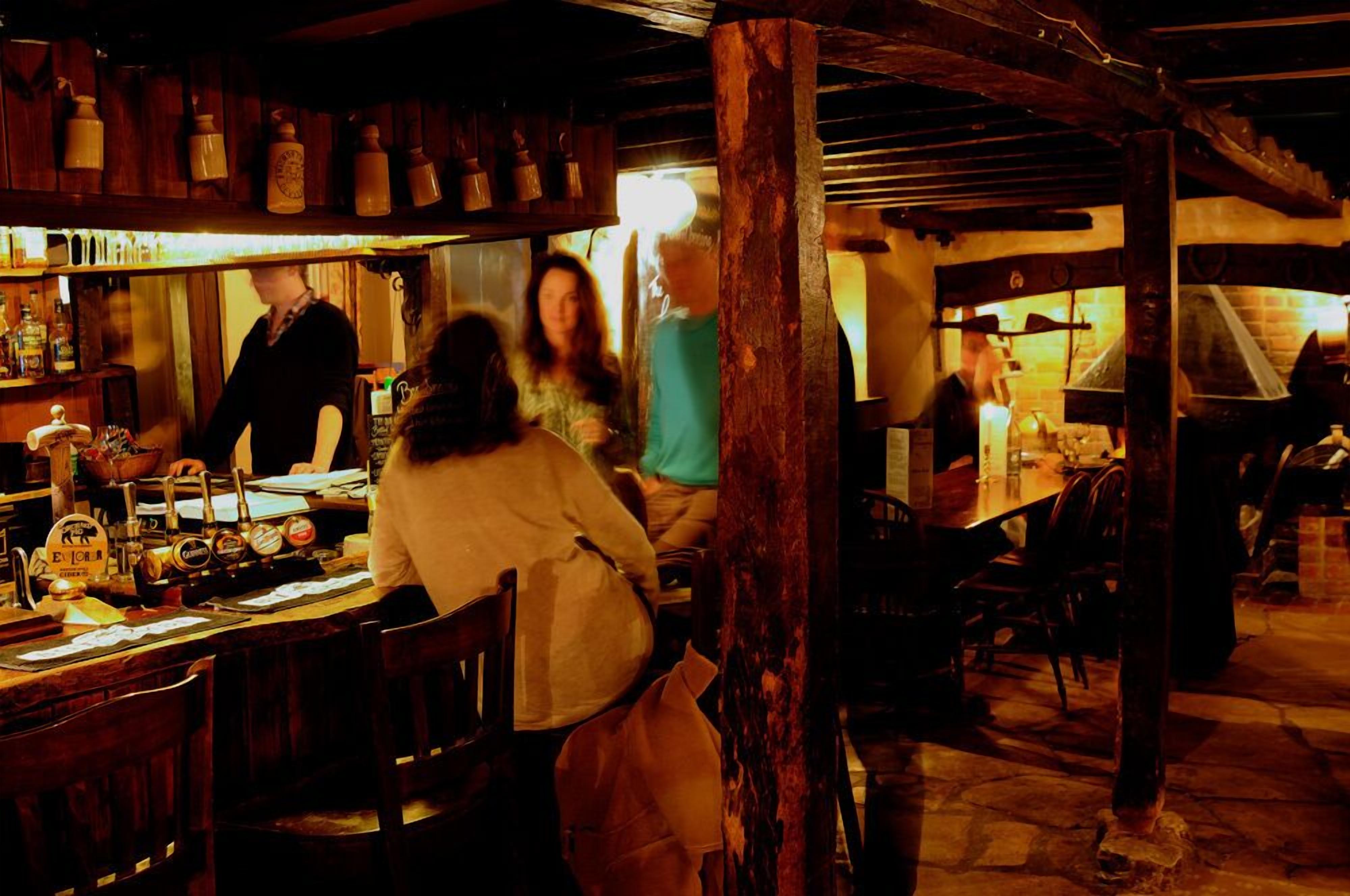 A rustic interior with wooden beams, a bar area with bottles, and a brick oven in the background.