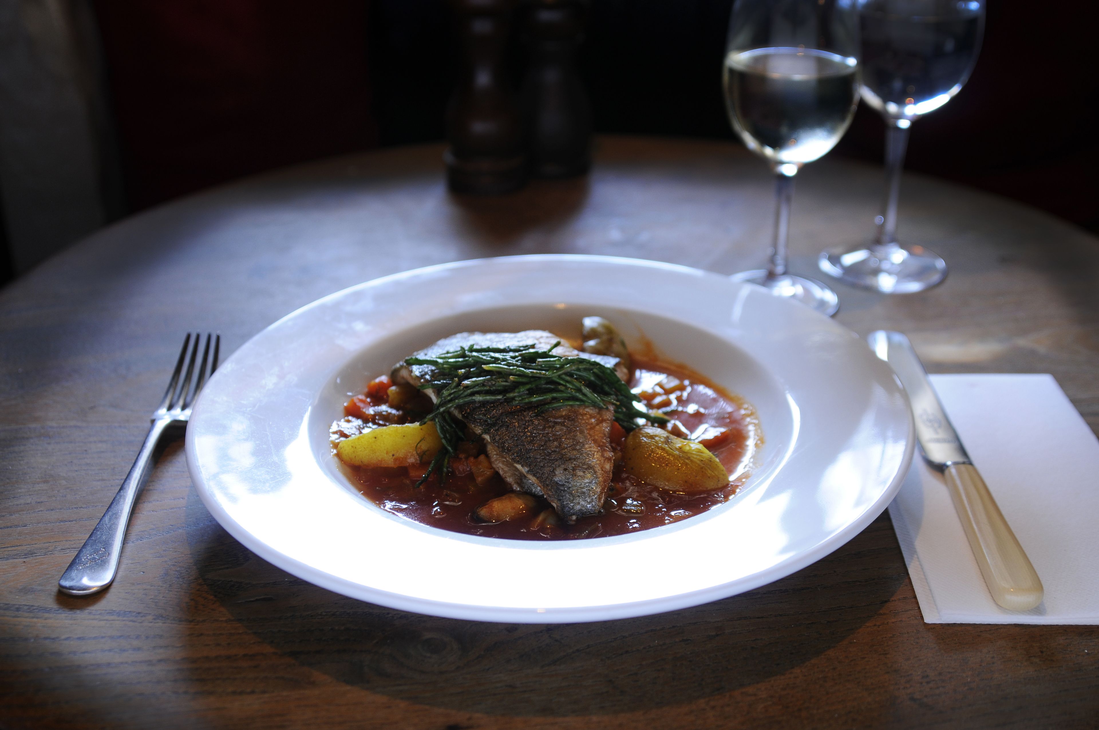 A plate of fish with vegetables and herbs, a fork, a knife, and two glasses of water on a wooden table.