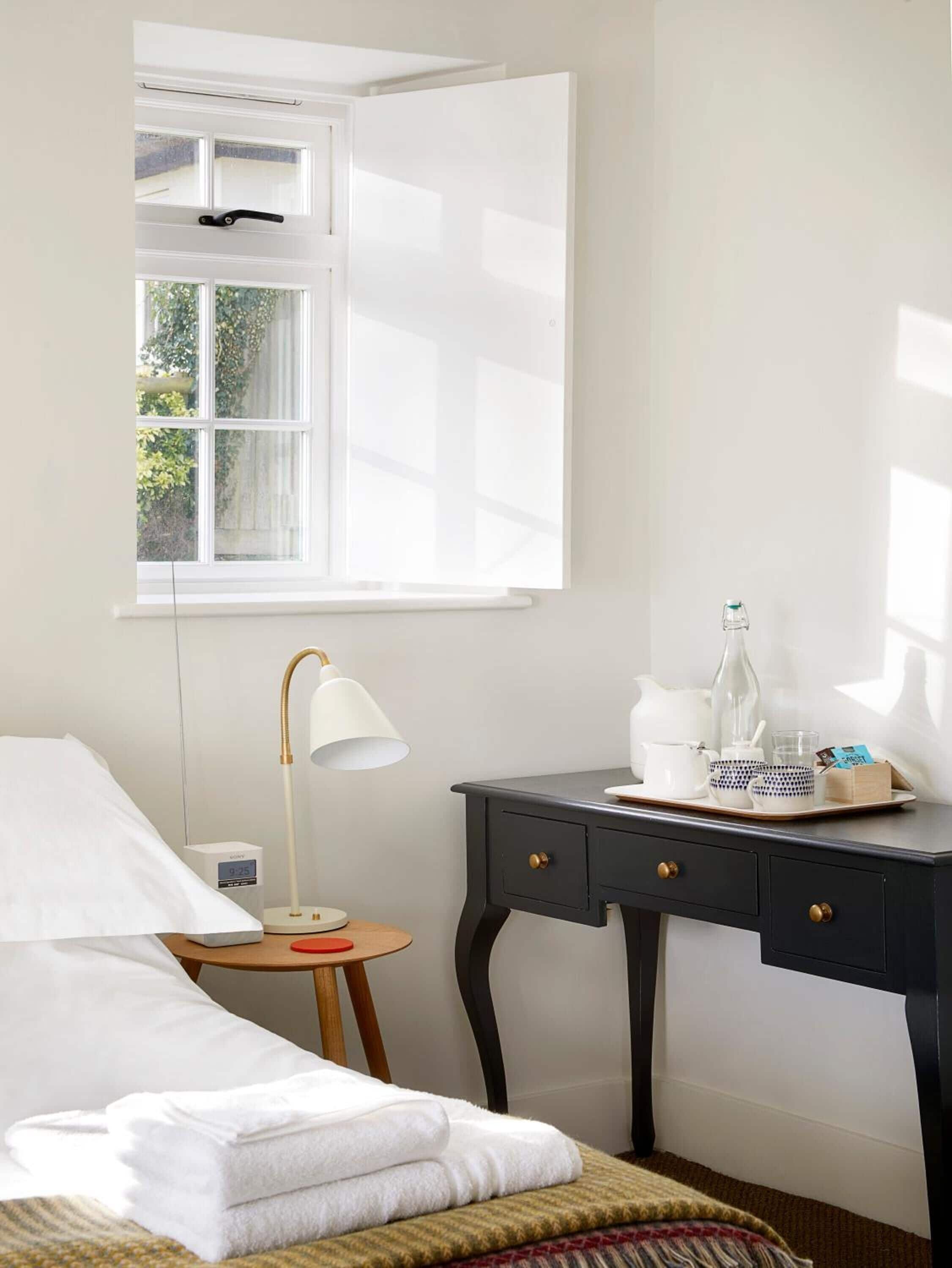 A hotel room with a bed, a bedside table with a lamp, a small table with a clock, and a window with a view of greenery.