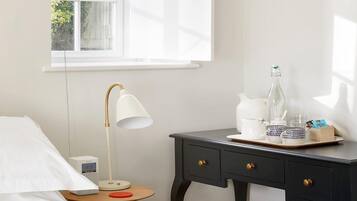 A hotel room with a bed, a bedside table with a lamp, a small table with a clock, and a window with a view of greenery.