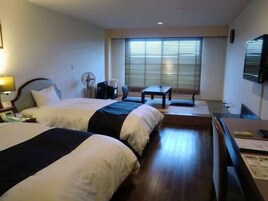Habitación de hotel con dos camas, un escritorio, una silla, un ventilador y una ventana con persianas.