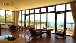 Un'ampia stanza con ampie finestre che offrono vista sul mare. Diverse tavole in legno e sedie in vimini disposte per mangiare.