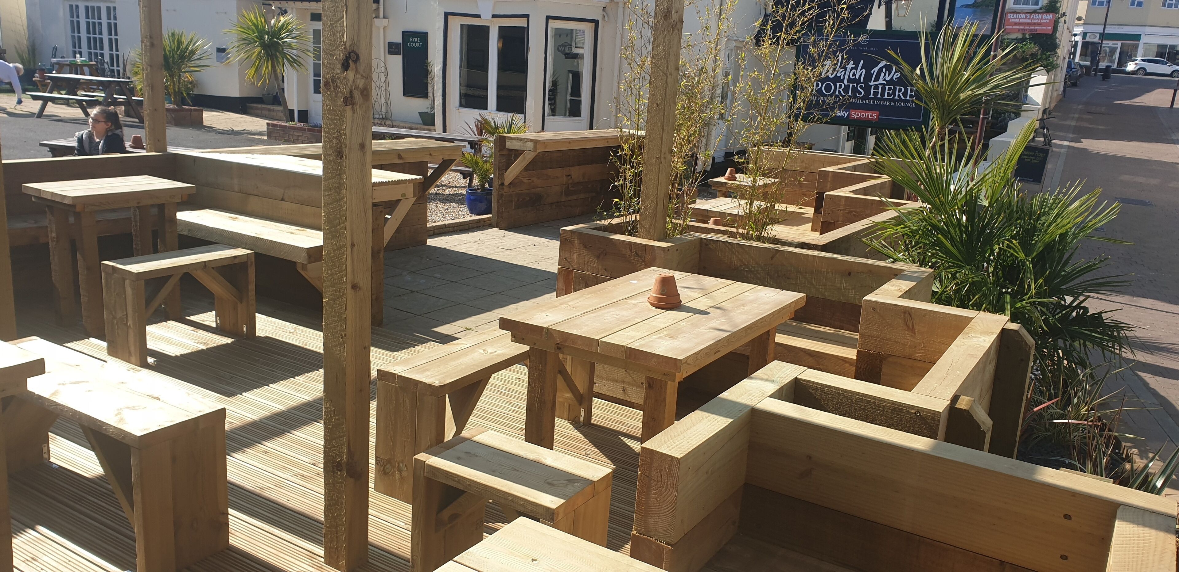 Un espacio al aire libre con mesas y bancos de madera, rodeado de plantas en macetas.