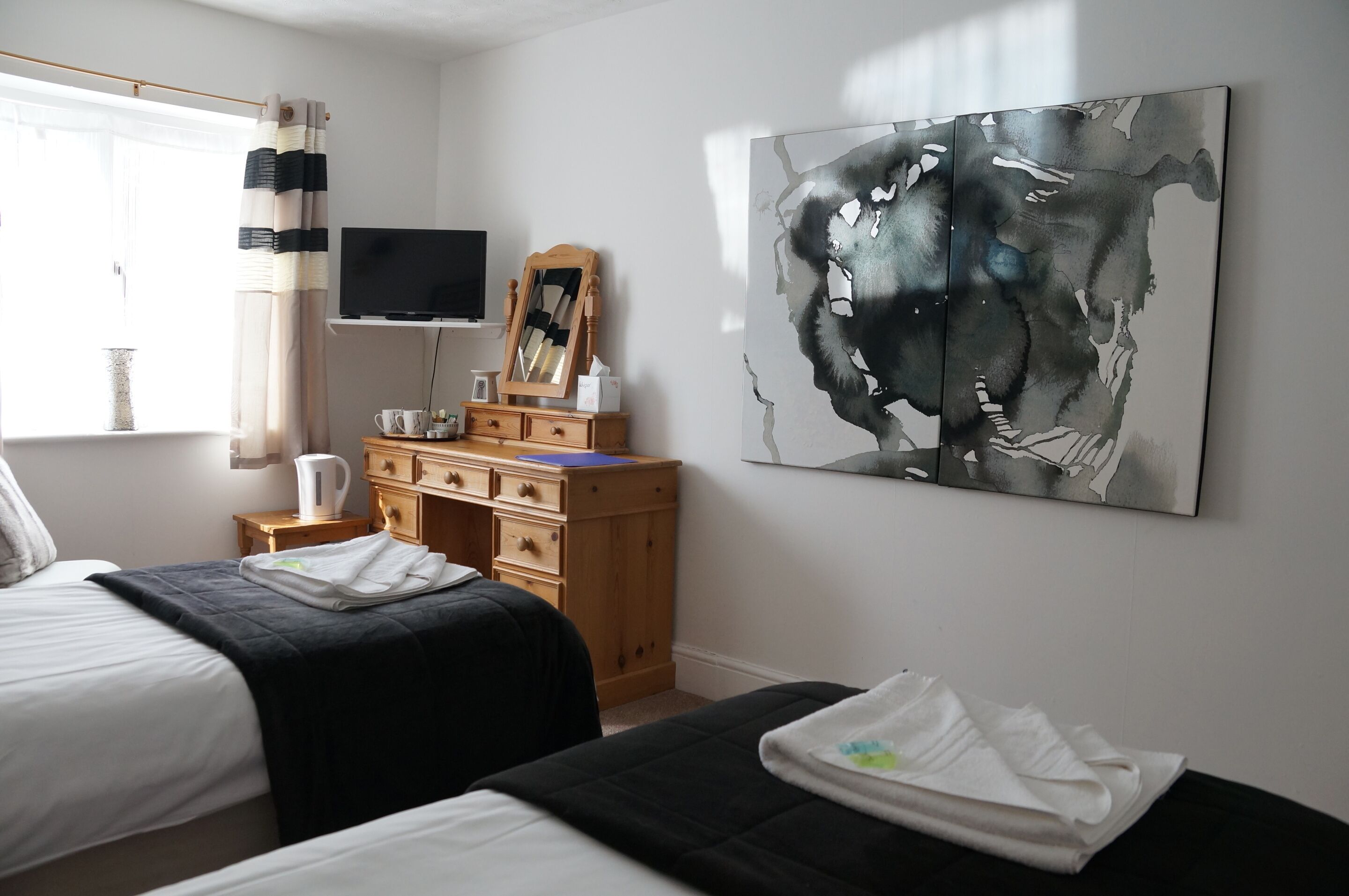 A bedroom with two beds, a wooden dresser, a flat-screen TV, a large abstract painting, and a window with curtains.