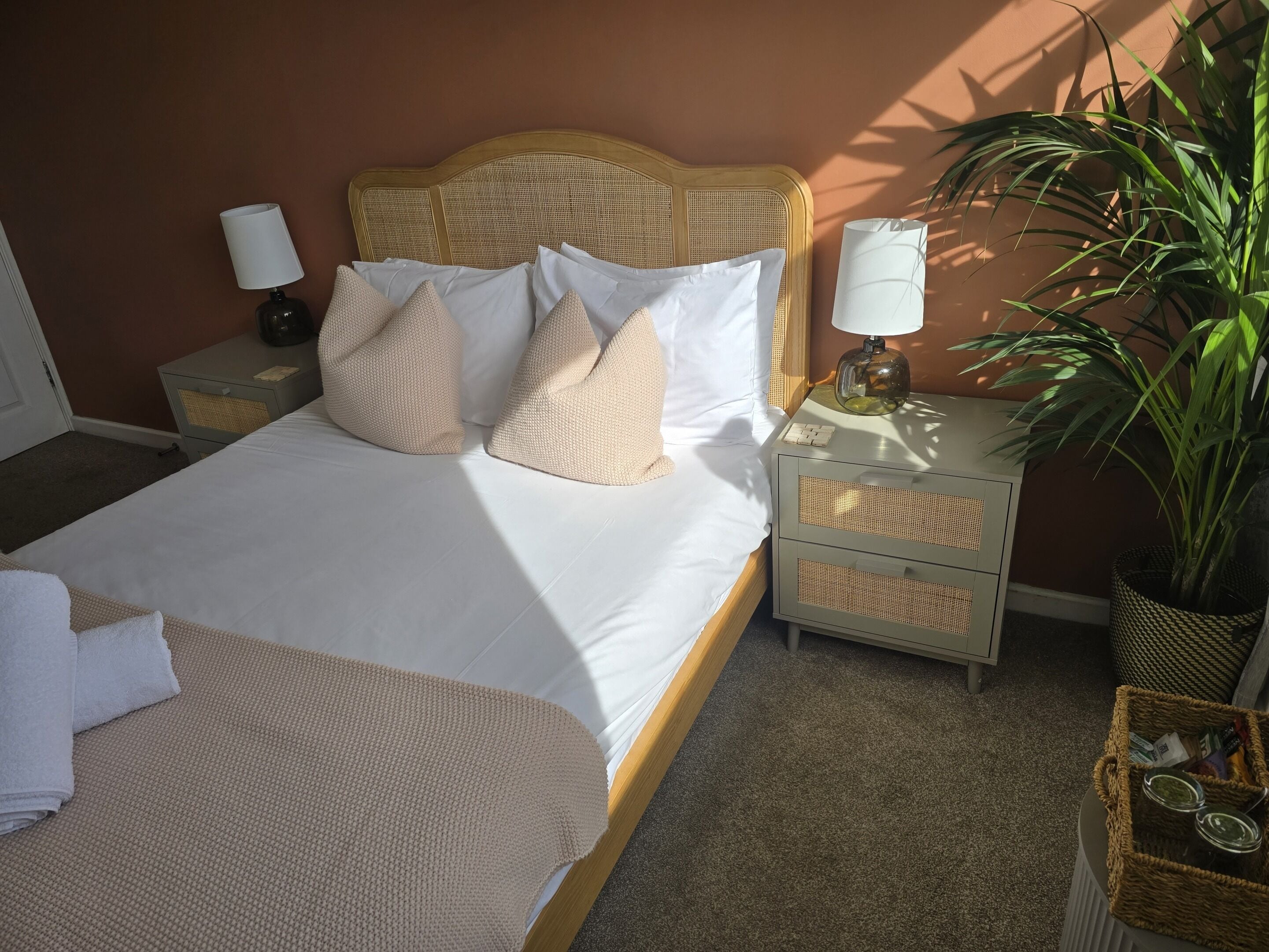 Un lit bien fait, avec du linge de lit blanc et des coussins beige, une lampe de chevet, une petite table avec une lampe et une plante en pot.