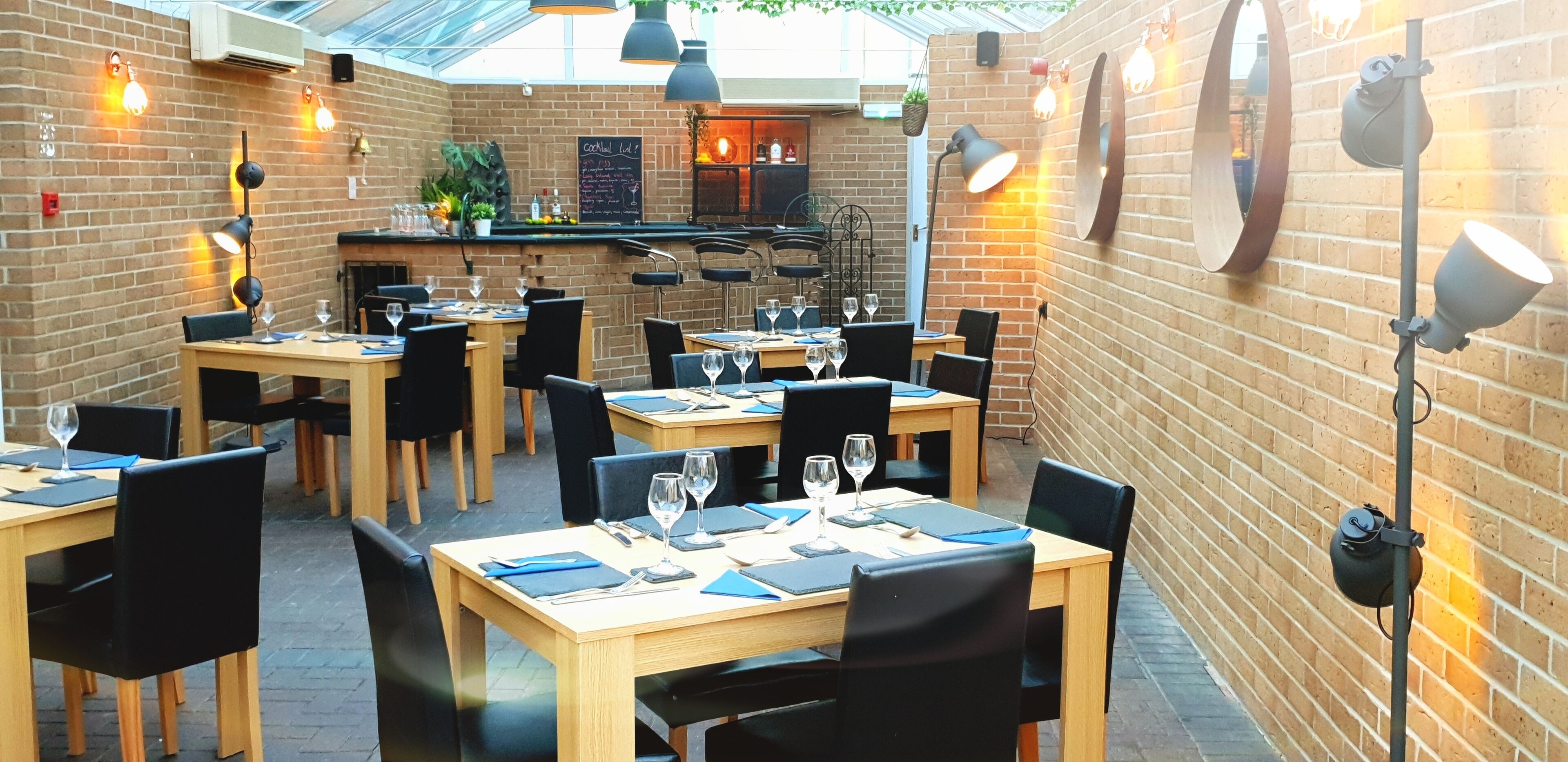 An outdoor restaurant with brick walls, wooden tables, black chairs, and hanging pendant lights.