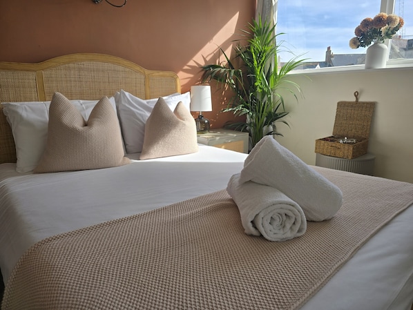 A neatly made bed with a rolled towel, a rattan headboard, and a potted plant beside the window. - Eyre Court Hotel (Seaton)