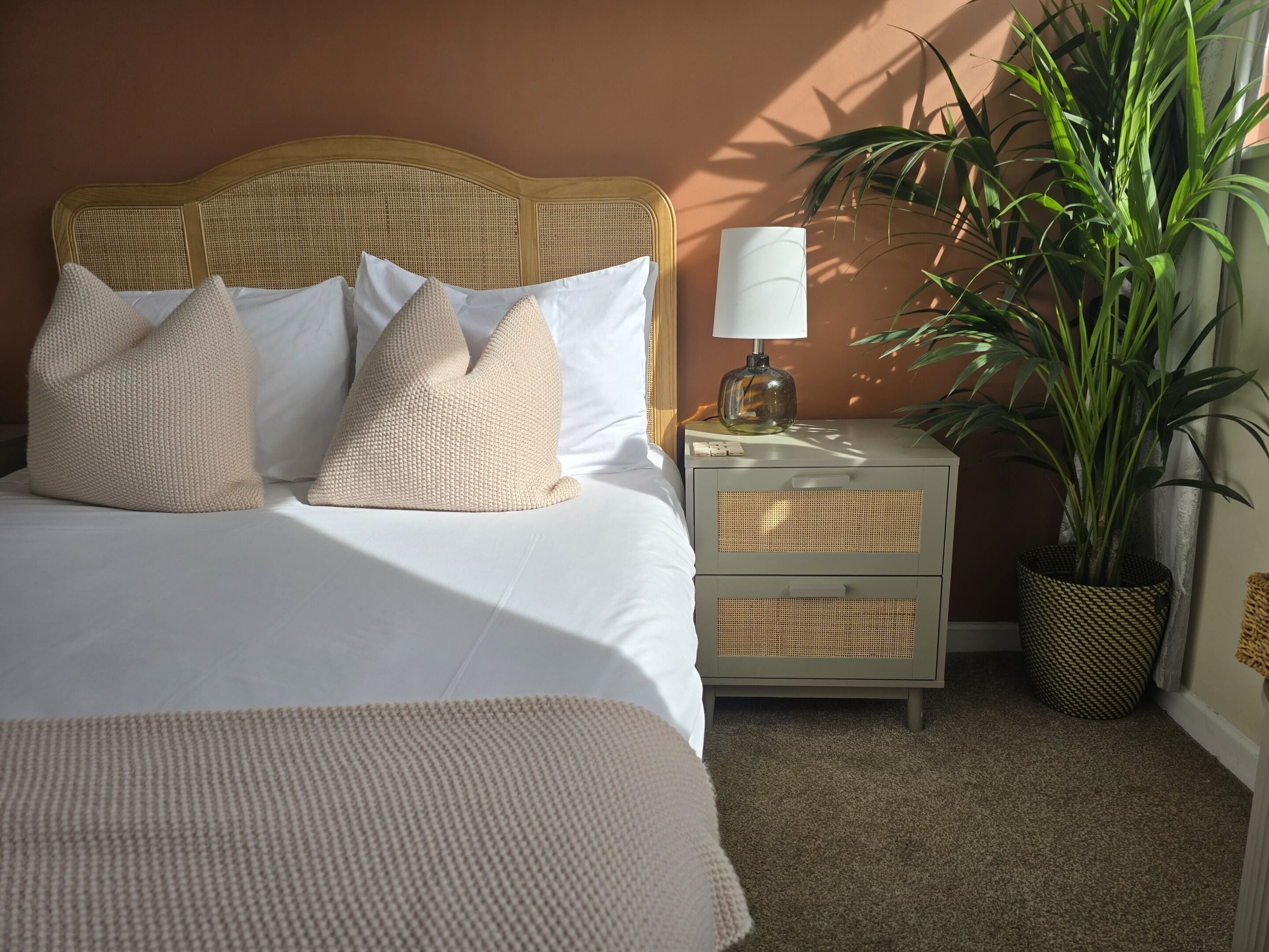 A neatly made bed with white linens and beige pillows, a bedside lamp, and a potted plant.