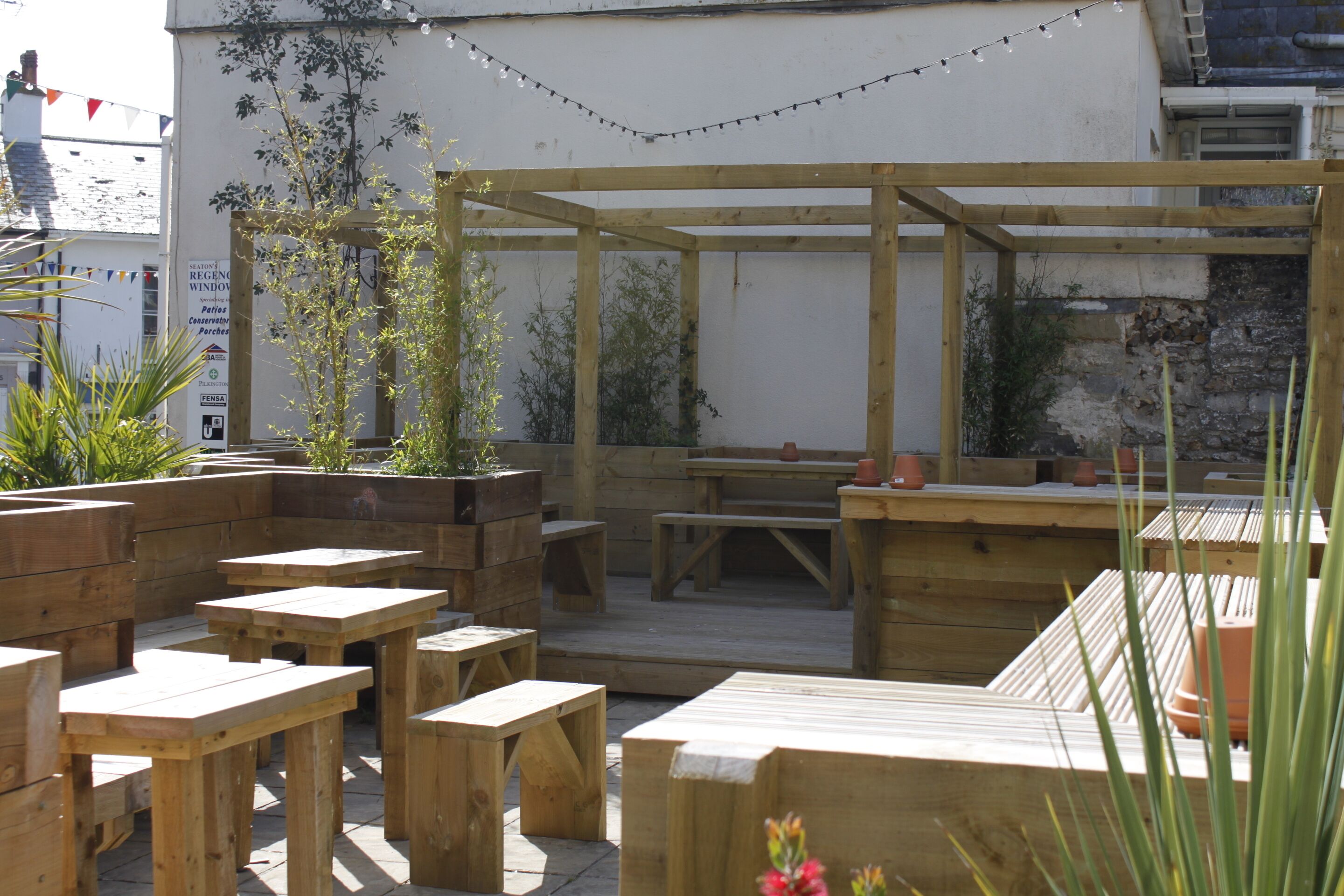 En udendørs terrasse med træborde og bænke, omgivet af plantekasser og lyskæder.