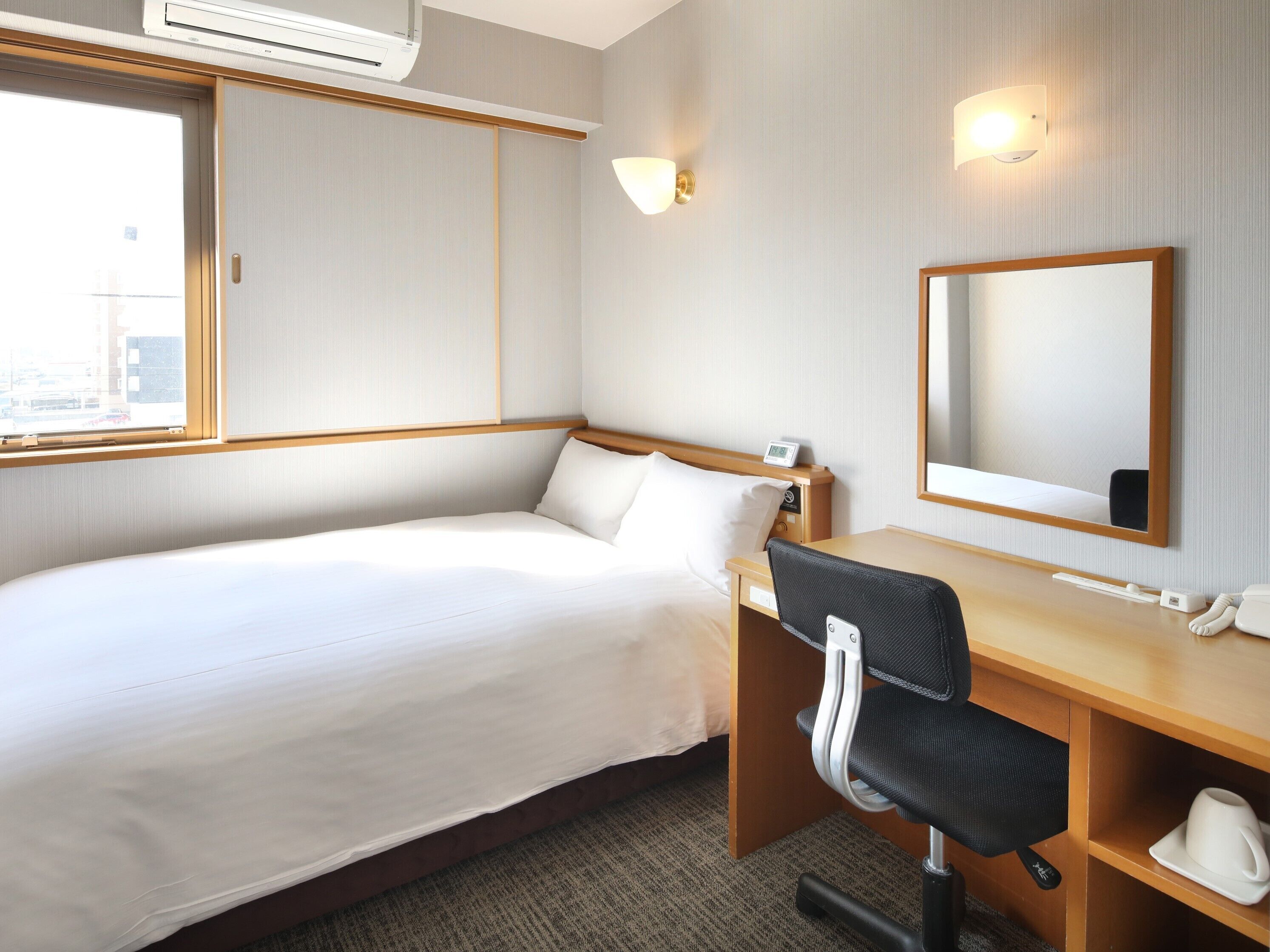 A hotel room with a bed, a desk, a chair, a mirror, and a bedside table with a phone and a cup.