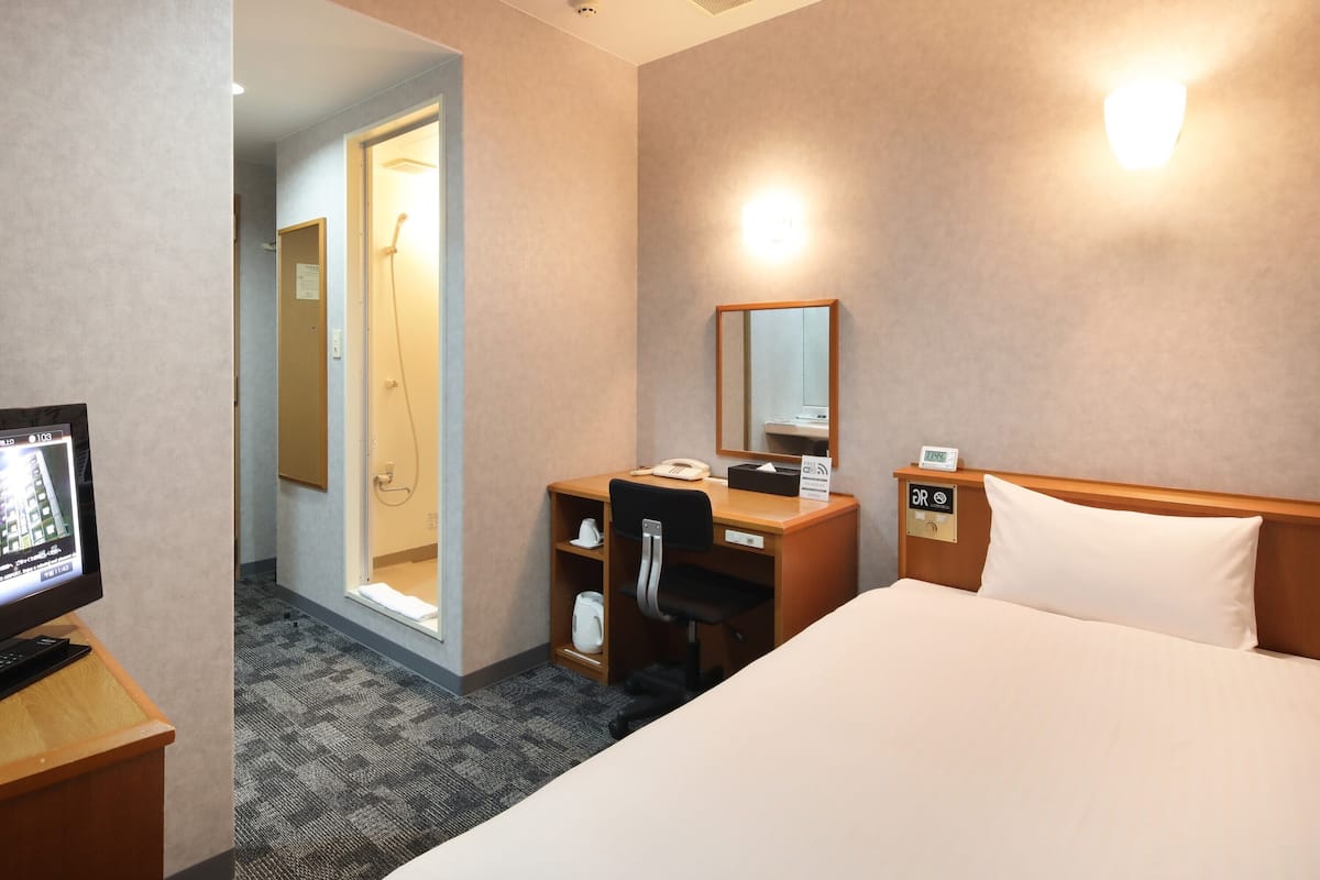 A hotel room with a bed, desk, chair, and a shower area.