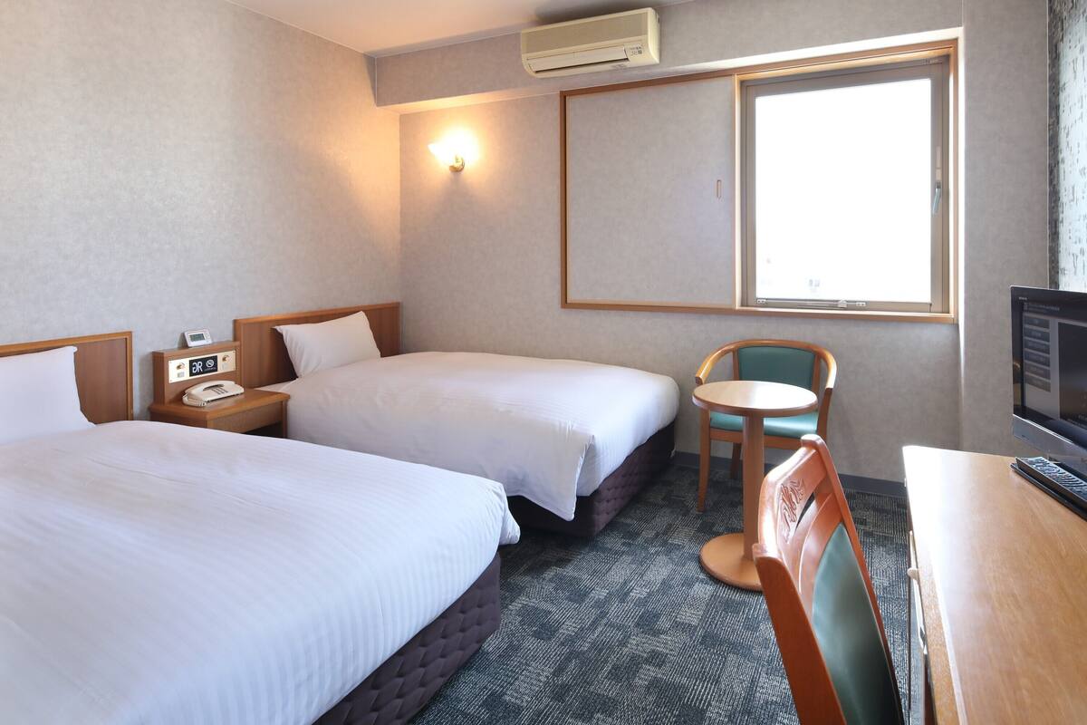 A hotel room with two beds, a desk, a chair, a TV, a phone, and an air conditioner.