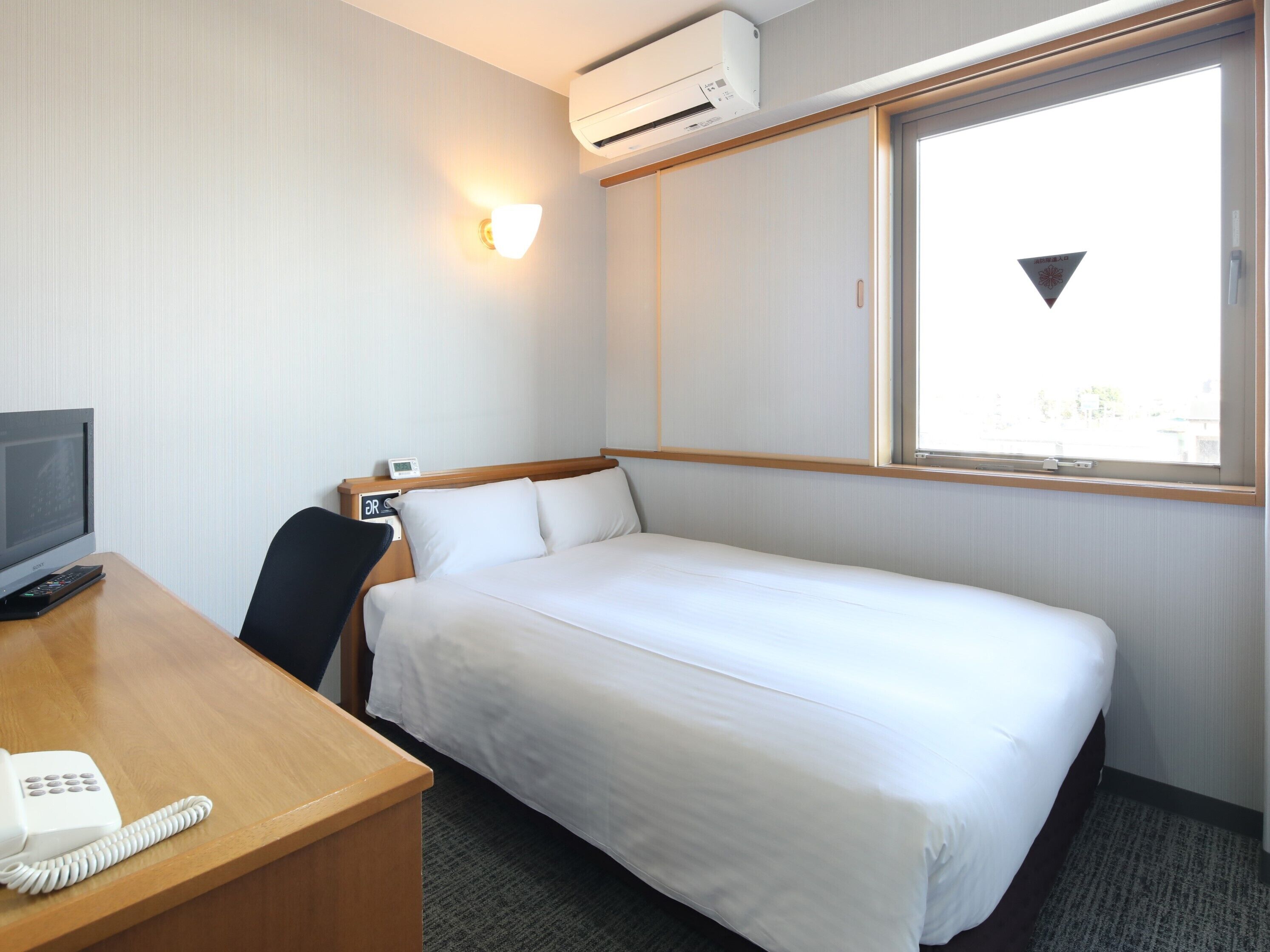 A hotel room with a single bed, a desk with a telephone, a TV, and an air conditioning unit.