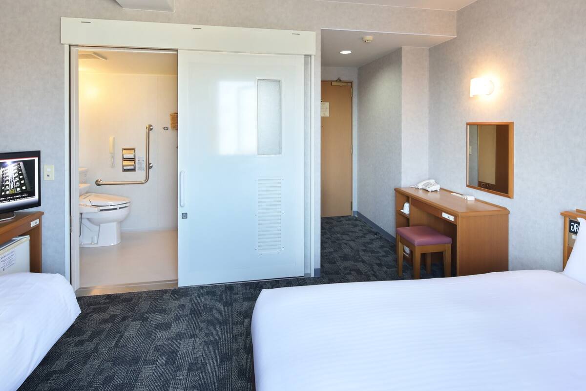 A hotel room with a bed, a desk with a chair, a bathroom with a toilet and sink, and a mirror on the wall.
