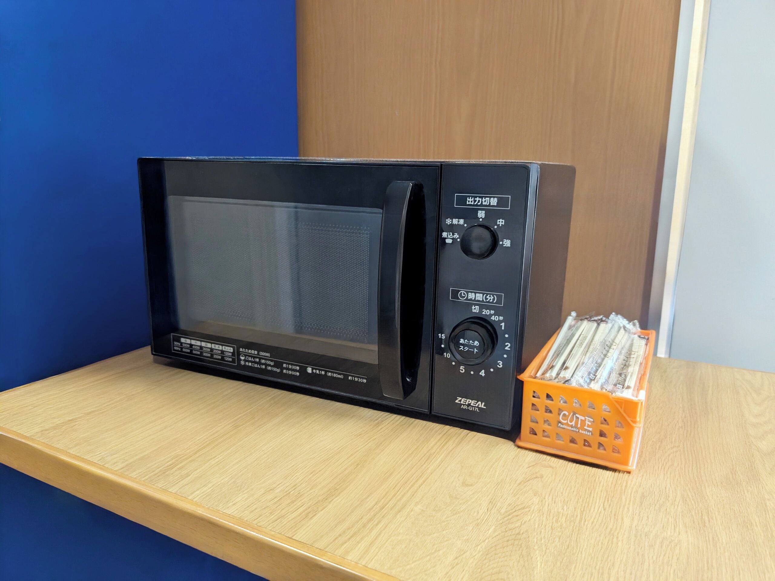 Un four à micro-ondes noir est posé sur une étagère en bois, avec un mur bleu et une porte en bois en arrière-plan.
