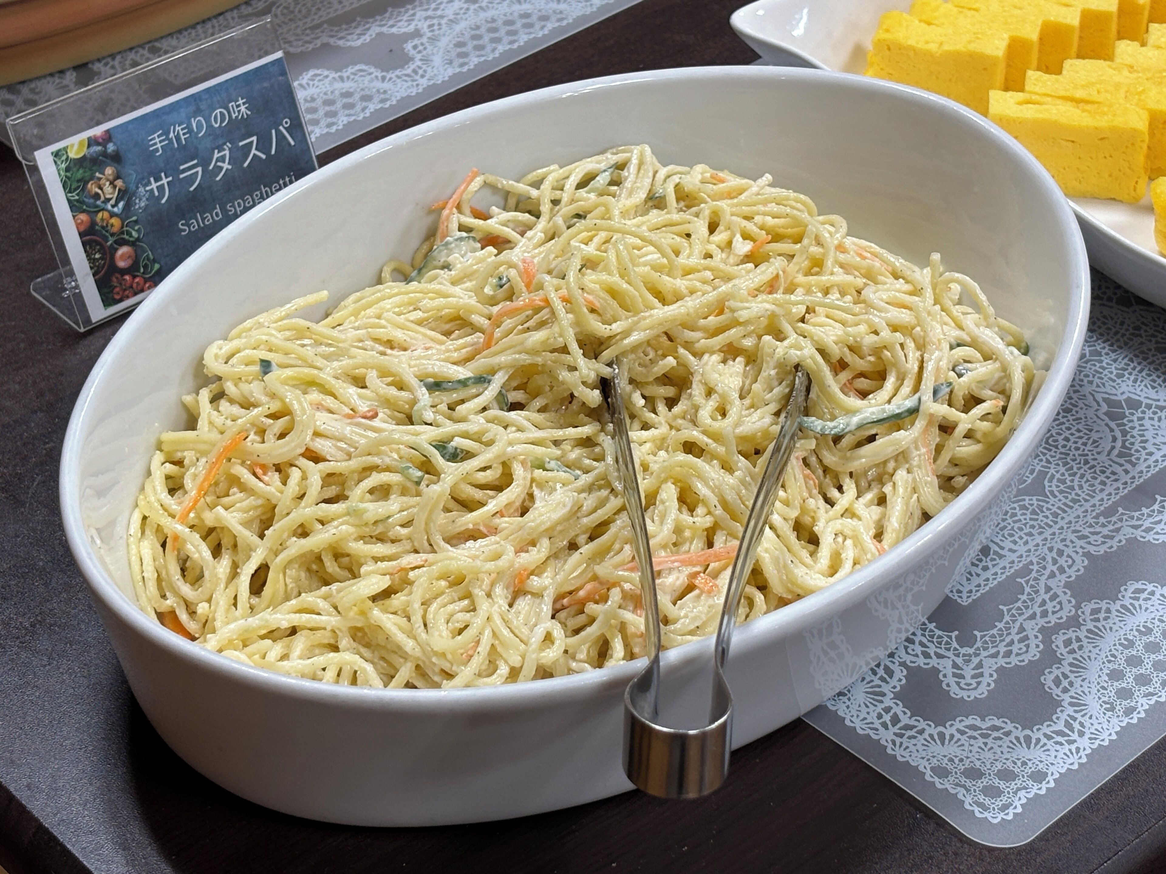 Un bol de spaghetti à la salade, accompagné d’une cuillère, posé sur une table recouverte d’une nappe en dentelle.
