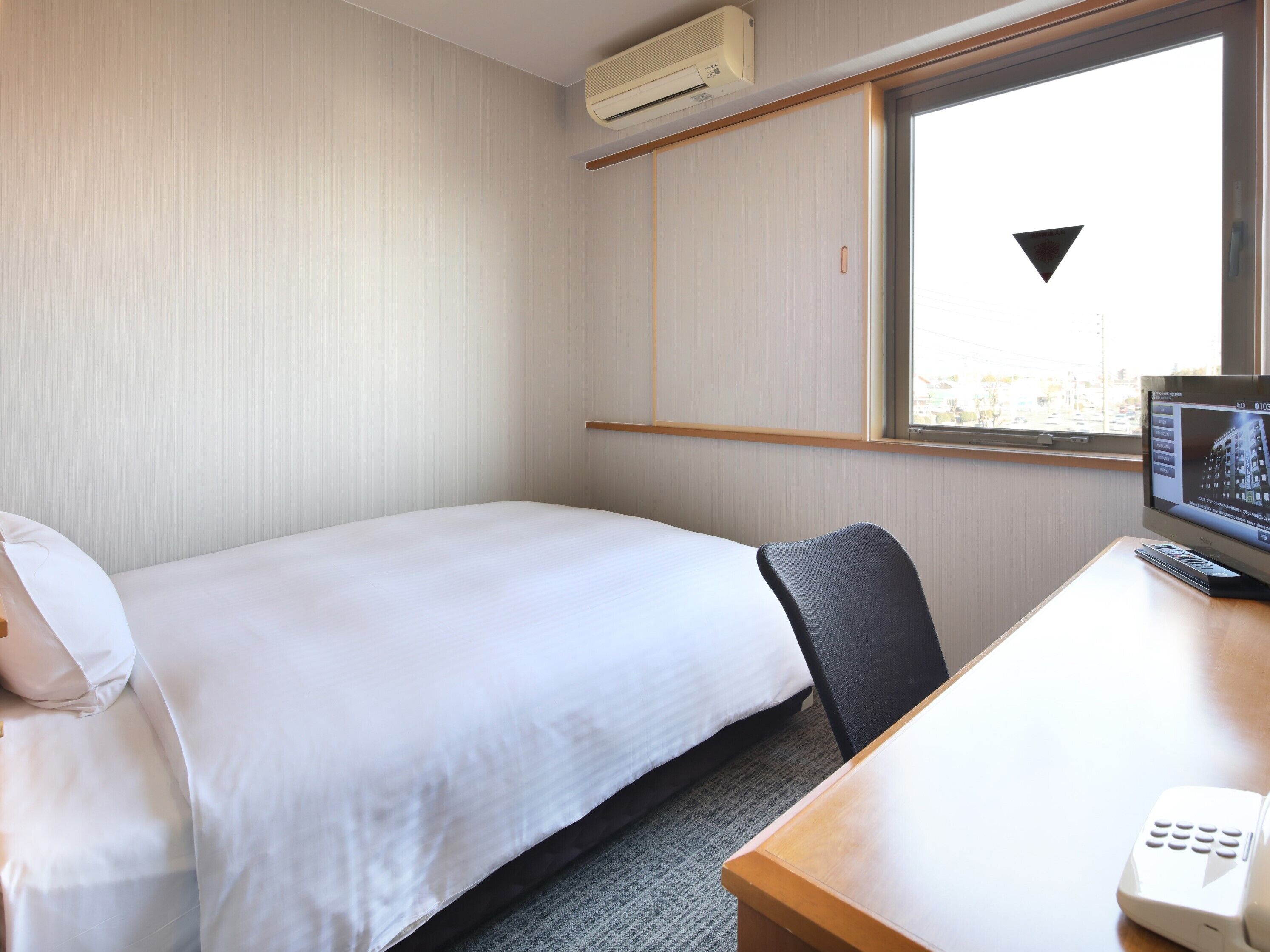 A hotel room with a bed, a desk with a computer monitor, a remote control, and a window with a view of buildings.